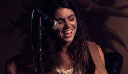 Sheera strums an acoustic guitar while standing in front of a microphone.